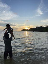 Rear view of dad and son standing at beach against sky during sunset