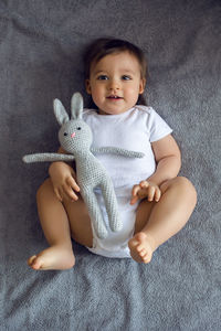 Very cheerful baby boy in white clothes lying on the bed with a knitted toy rabbit