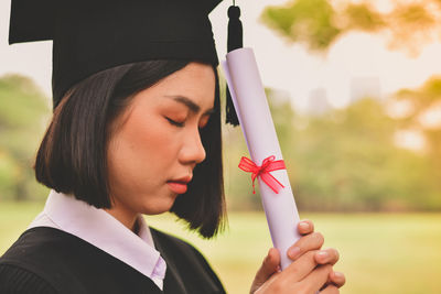 Close-up of young woman in graduation gown praying while standing at park