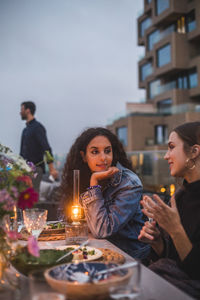 Smiling female friends talking while sitting on building terrace during sunset