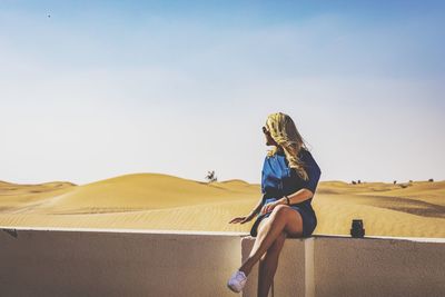 Woman sitting on retaining wall at desert against sky