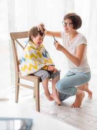 Mother cuts her son's hair by herself. little boy sits, covered with cloth,holds pair of scissors. 