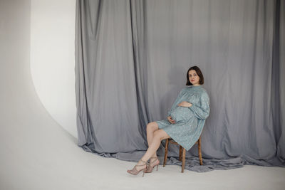 Pregnant woman sitting on chair against gray backdrop