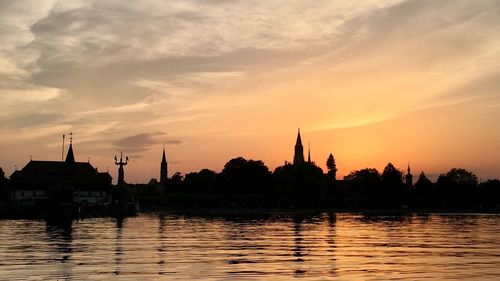 Silhouette buildings by lake against sky during sunset