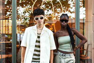 Young diverse man and woman in trendy clothes and sunglasses standing against reflective glass wall in daytime on city street