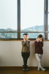 Full length of cute siblings standing with head in hands against window