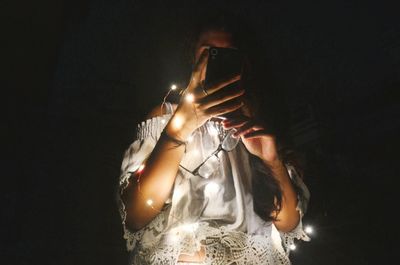 Low angle view of woman with illuminated string lights using mobile phone in darkroom at home