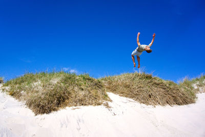 Low angle view of young woman jumping on beach against clear blue sky