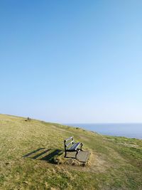 Empty bench on field by sea against clear blue sky