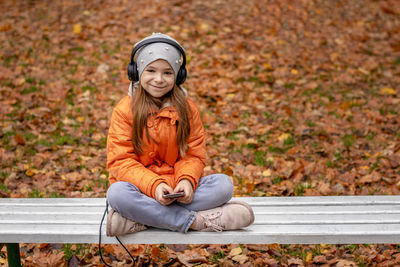 A girl listens to music on headphones in an autumn park while sitting on a white bench.