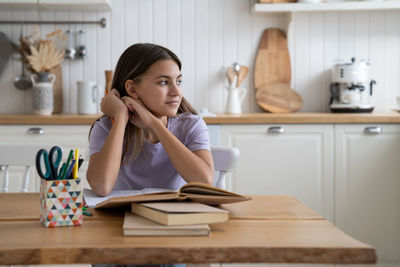 Dreamy optimistic teenage girl sits in appartment at kitchen table with books and looking to side