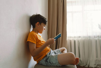 A cute boy wearing an orange t-shirt is sitting on a soft ottoman reading a book