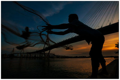 Silhouette fisherman throwing fishing net by bridge against sky during sunset