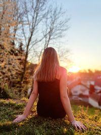 Rear view of woman sitting on field against sky during sunset