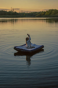 Young woman sits on a mattress on the water with her hands up during sunset