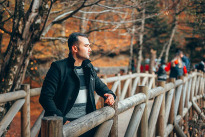 Young man looking away while standing by railing against trees