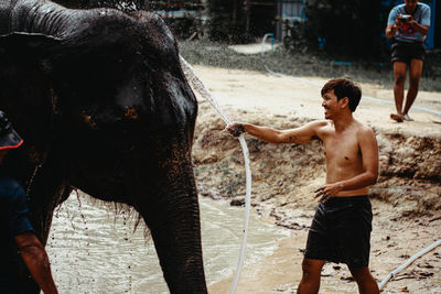 Shirtless man washing elephant with water pipe while standing in lake