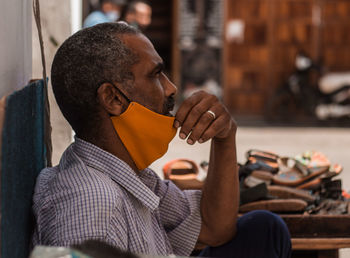 Thoughtful cobbler wearing mask sitting on street