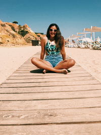 Smiling young woman sitting with cross-legged on boardwalk at beach during sunny day