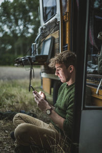 Side view of man sitting cross-legged while using smart phone against motor home during camping
