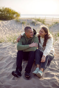 Husband and wife in hoodies and sneakers sitting on a sandy beach by the sea in autumn