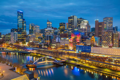 Illuminated modern buildings by river against sky in city