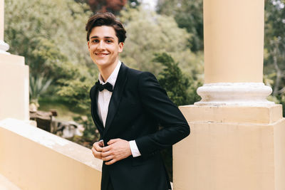 A beautiful young man, the groom in an elegant wedding suit, stands posing in the city's old park