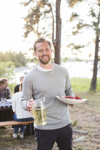 Portrait of happy man holding beer jug and strawberries with friends sitting at picnic table in background