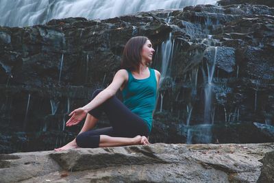 Young woman performing yoga on rock against waterfall