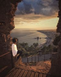 Woman sitting by sea against sky during sunset