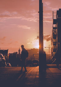Silhouette man walking on street by buildings in city sky during sunset