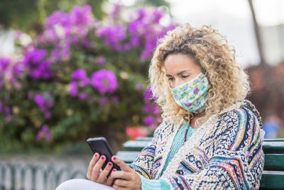 Smiling woman wearing mask using mobile phone while sitting outdoors