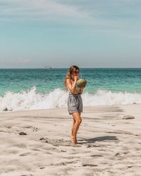 Full length of woman drinking coconut water while standing at beach against sky