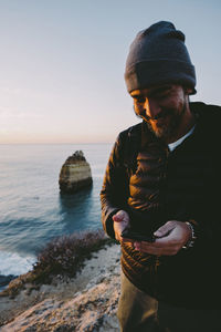 Man using mobile phone while standing on rock formation against sky during sunset
