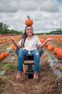 Smiling young woman balancing pumpkin on head while sitting in wheelbarrow at field against sky