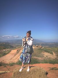 Full length of woman with puppy standing on hay bale by landscape against blue sky