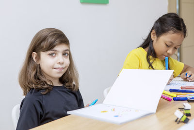 Portrait of girl sitting with friend while holding felt tip pen in classroom at school