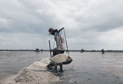 Side view of man carrying salt in baskets on shore against cloudy sky