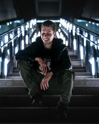 Low angle view of smiling young man sitting on illuminated steps