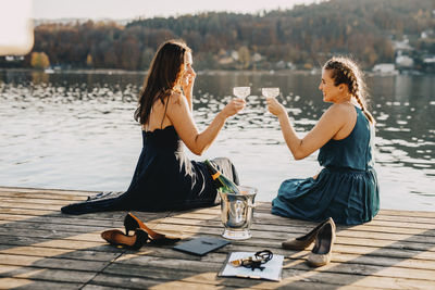 Female event planners toasting champagne while sitting on jetty over lake