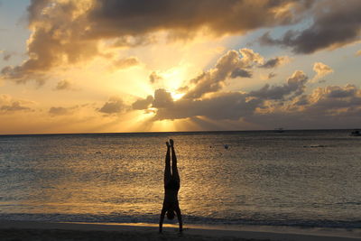 Silhouette person doing handstand on shore at beach against cloudy sky during sunset