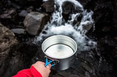 Cropped hand of man holding metal container against waterfall at campsite