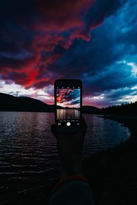 Man photographing illuminated smart phone against sky at sunset