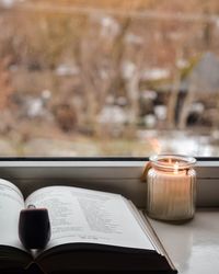 Close-up of open book by illuminated candle on window