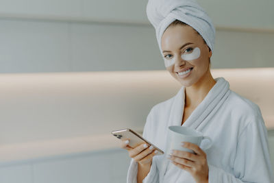 Portrait of smiling young woman with medical eye patches holding coffee cup and mobile phone at home