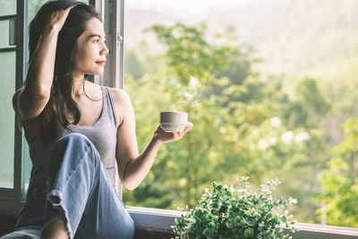 Young woman looking away while sitting on window