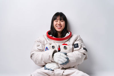 Young asian female in spacesuit looking at camera with smile while sitting near gray wall during space mission