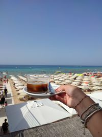 Cropped image of hand holding drink at beach against clear sky