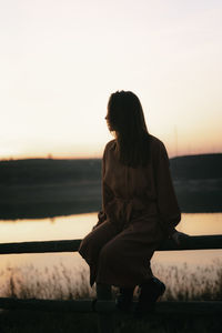 Young woman sitting on railing looking at lake against sky during sunset