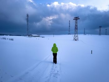 Woman with green hooded jacket walking down the snow towards transmission towers on a cloudy day